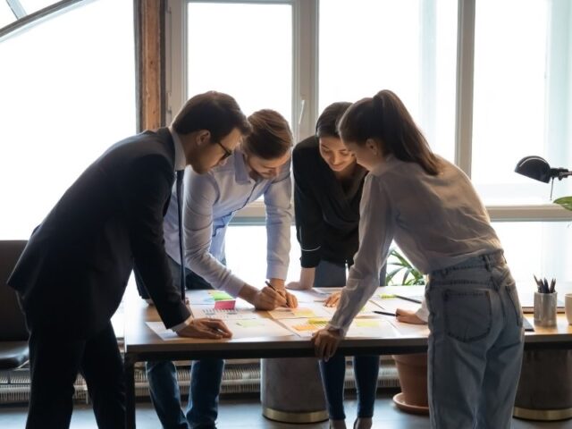 Four colleagues brainstorm around a table covered with sticky notes and papers in a bright office space.