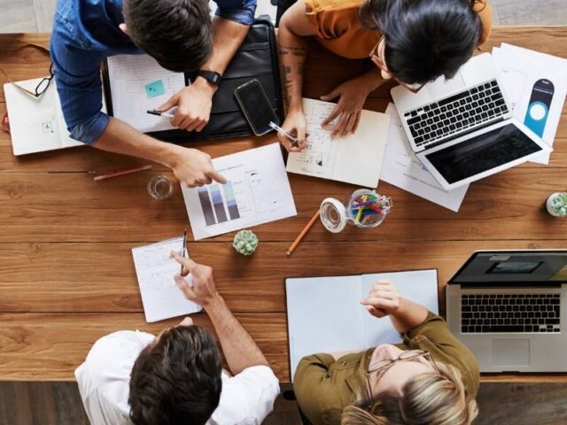 Group of four professionals working around a wooden table with laptops, charts, and notes.